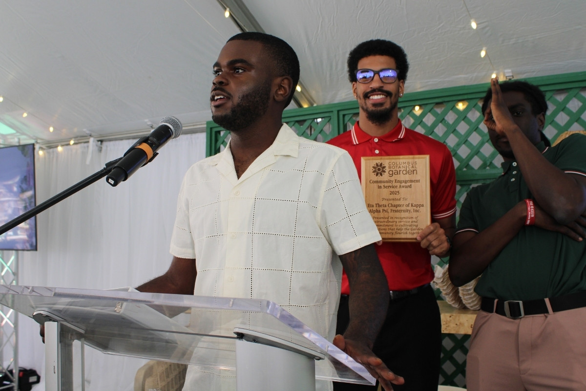 A young man in a white button-down shirt speaks at a podium during an event. Behind him, another man in a red polo shirt smiles while holding a wooden plaque from the Columbus Botanical Garden. The plaque reads: "Community Engagement in Service Award 2025, Presented To: Eta Theta Chapter of Kappa Alpha Psi, Fraternity, Inc." A third man in a green shirt stands to the right under a white event tent with string lights.