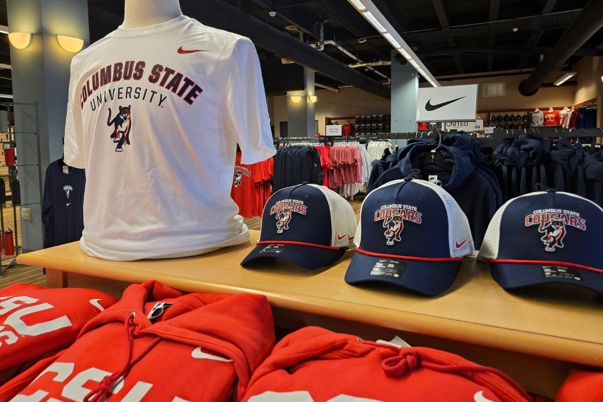 A selection of Nike-branded Columbus State University Cougars merchandise, including t-shirts, trucker hats, and red hooded sweatshirts displayed on clothing racks and counters.