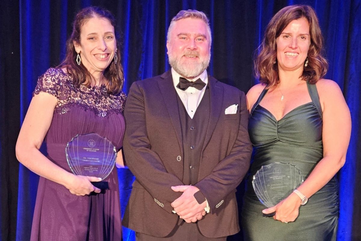 Three people in formal evening wear pose together in front of a dark blue curtain backdrop. The woman on the left in a purple lace-trimmed gown and the woman on the right in a dark green gown each hold fan-shaped glass University System of Georgia awards. The man in the center wears a dark tuxedo with a bowtie and pocket square.