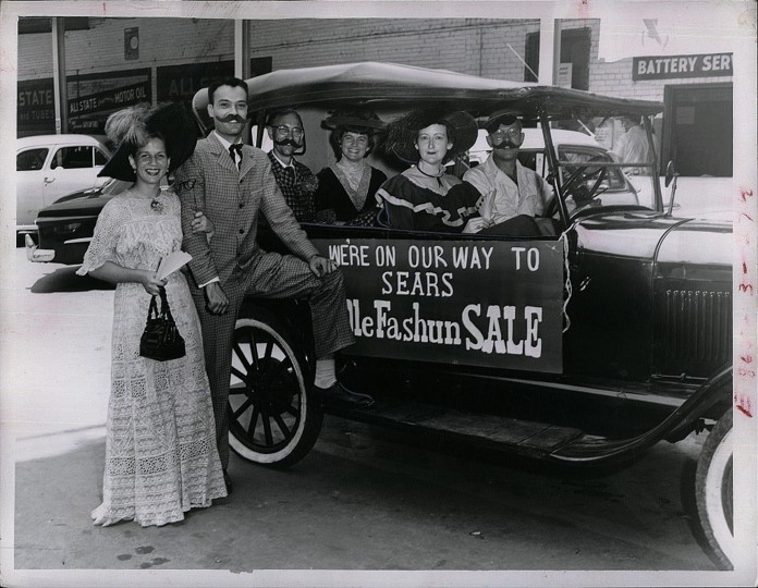 Black and white photo of people riding in an antique car with a sign on it that reads: we're on our way to Sears. Ole Fashun SALE