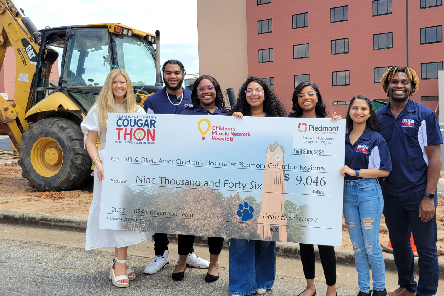 Group of people posed in front of a construction site holding a $9,046 "big check" to Bill and Olivia Amos Children's Hospital at Piedmont Columbus Regional