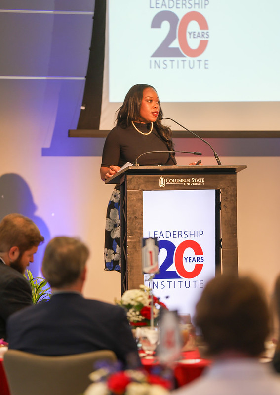 Breana Jones stands at a podium smiling and making remarks to the anniversary luncheon audience.