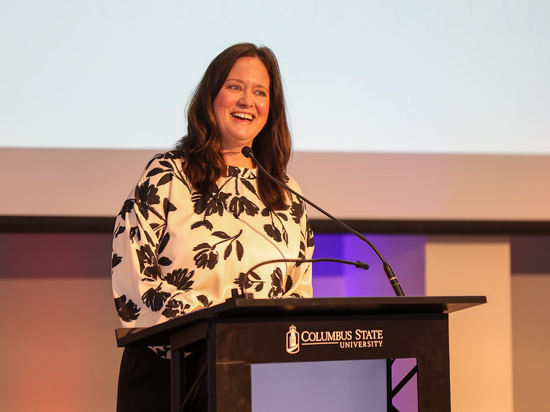 Shana Young stands at a podium smiling and making remarks to the anniversary luncheon audience.