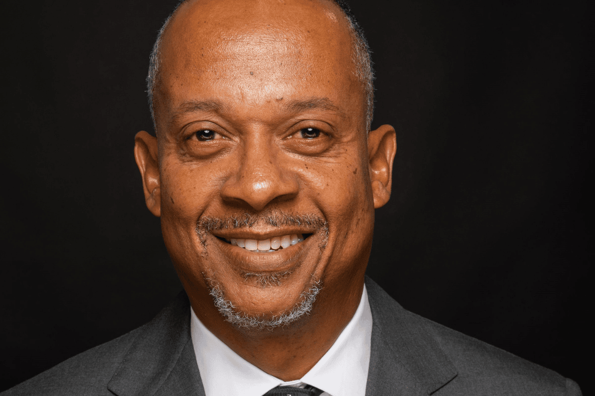 A close-up headshot of a smiling middle-aged Black man with a shaved head and a salt-and-pepper goatee. He is wearing a dark gray suit jacket, a white dress shirt, and a dark patterned tie against a solid black background.
