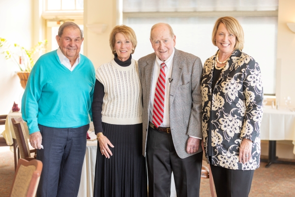A group of four smiling adults stands together in a well-lit indoor dining or event space. From left to right, Jimmy Yancey, Pamela Thayer, John Thayer Sr., Stuart Rayfield. The group is positioned in front of a white-clothed table in a room with large windows, suggesting a professional or celebratory gathering.