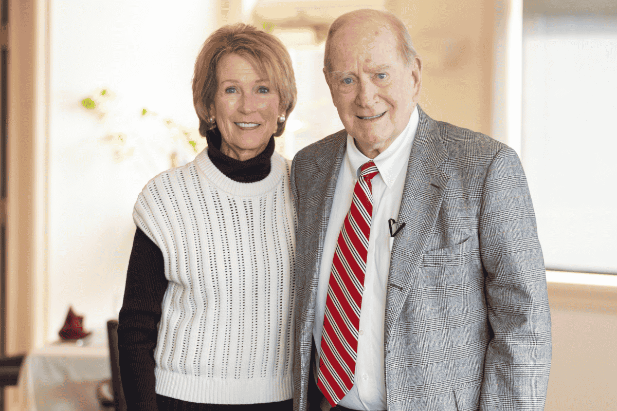 Pamela and John Thayer stand side-by-side, smiling warmly at the camera in a brightly lit indoor setting. The woman, on the left, is wearing a black turtleneck layered under a white sleeveless knit sweater. The man, on the right, wears a grey patterned blazer over a white button-down shirt and a bold red-and-white striped tie. The background is softly blurred, showing hints of a white table and a window, suggesting a comfortable, formal, or celebratory environment.