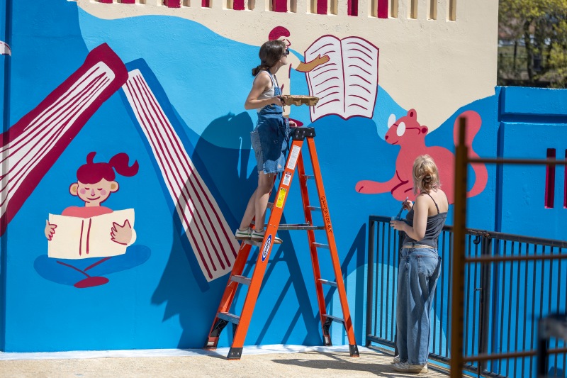 Cora Pearl James standing on a ladder and an assistant on the ground painting the mural.