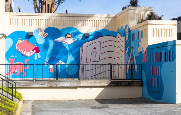 A blue and white mural on a Columbus State University campus wall featuring illustrated books, figures, and the words: excellence, creativity, and sustainability.