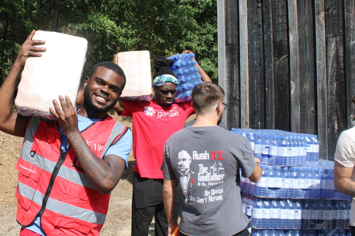 A group of young men carrying large cases of bottled water near a wooden building. In the foreground, a smiling man in a red "Community Service" vest with the Greek letters "K A Ψ" carries a case on his shoulder. Behind him, another man in a red "Columbus Botanical Garden" t-shirt carries two cases. A third man's back is to the camera, wearing a grey t-shirt that says "Rush K Σ" and features a parody of The Godfather logo with the text "An Offer You Can't Refuse." Multiple pallets of blue bottled water are stacked to the right.