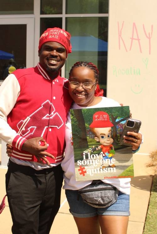 A smiling young man and woman stand together outdoors. The man wears a red and white letterman jacket and a red durag. The woman holds a poster featuring a Jimmy Neutron character wearing a Kappa Alpha Psi hat with text that reads, 'I love someone with autism.' In the background, 'KAY' is written on a wall in pink chalk.