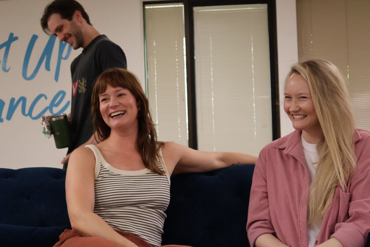 A candid shot of cast members during a break in a studio. The individuals are laughing and smiling together in a relaxed environment. Partially visible on the wall is a sign that reads 'Dance'