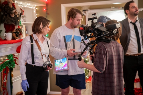 A film crew works on a decorated holiday set. On the left, an actor wearing elf ears, a white shirt with black suspenders, and blue latex gloves holds a camera. In the center and right, crew members manage a professional camera rig and a tablet. A man in a suit and tie stands in the background near a mantle decorated with Christmas greenery and lights.