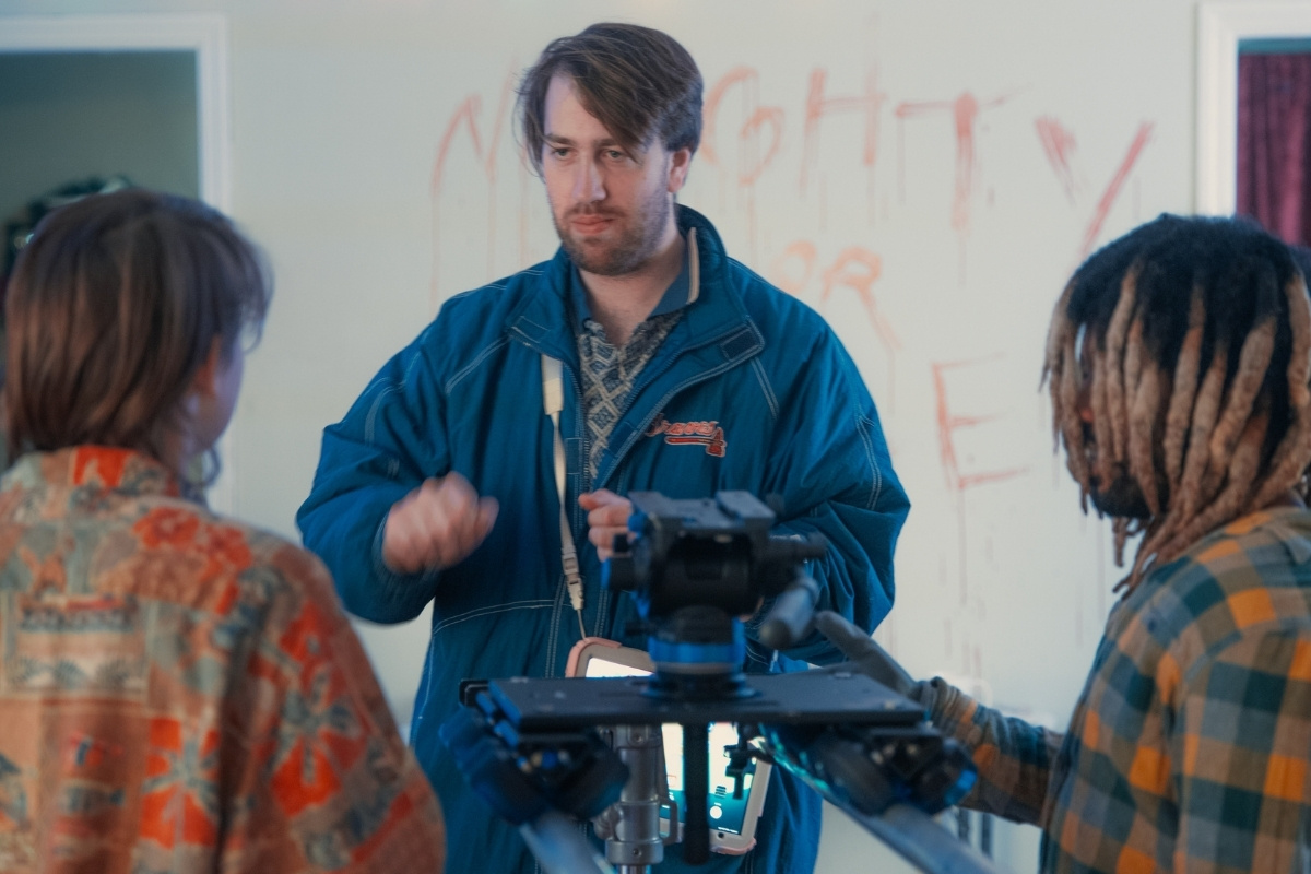 A behind-the-scenes look at a film set. A man in a blue jacket stands behind a camera rig, gesturing while speaking to two other individuals whose backs are to the camera. In the background, a white wall features large, stylized text scrawled in red paint that appears to read 