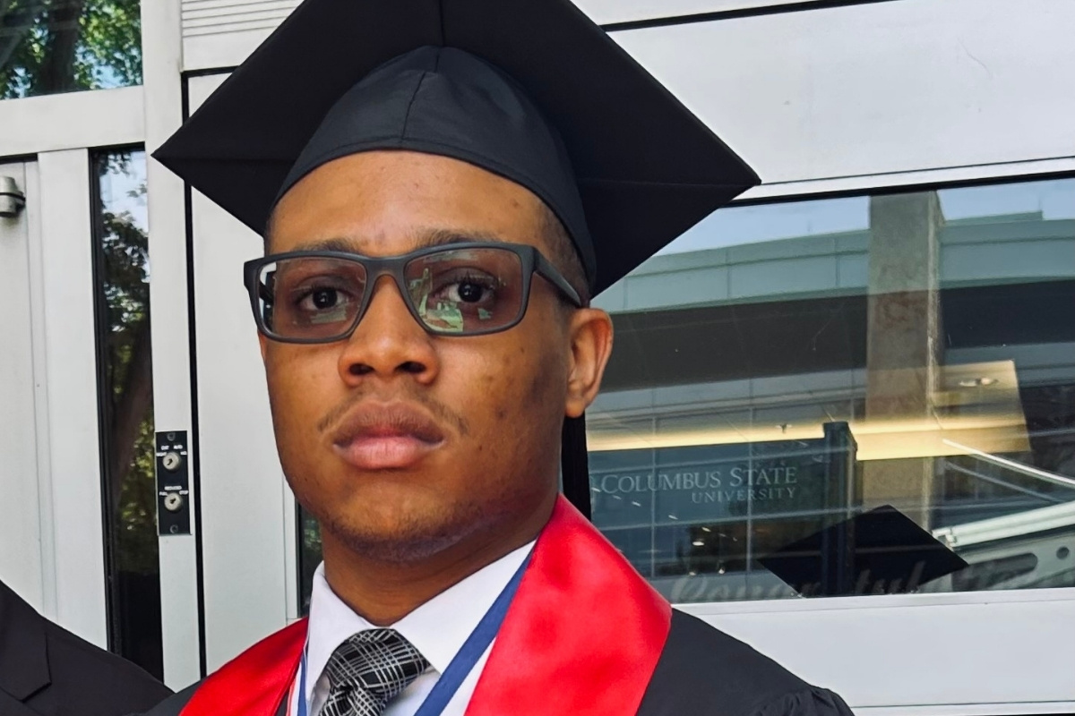 A close-up portrait of a male graduate with a serious expression, wearing a black graduation cap, glasses, a tie, and a red stole over his gown. A glass door behind him reflects a building with text that reads 