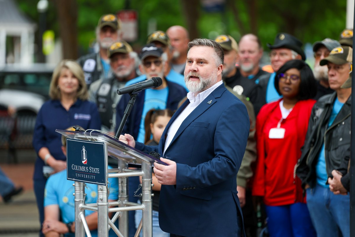 A speaker addresses a crowd from a Columbus State University podium during an outdoor event. He is wearing a dark blue blazer over a white shirt. The background shows several attendees listening attentively.