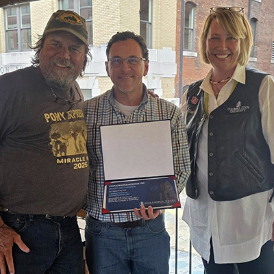 Three people stand smiling for a photo together on an outdoor wooden restaurant deck or patio under string lights. On the left, a man wearing a brown "Pony Express Miracle Ride 2025" T-shirt, jeans, and a baseball cap stands next to a man in the center wearing glasses, a plaid button-down shirt, and jeans, who is holding open an award certificate folder. On the right, a woman with blonde hair wearing a white shirt and a black leather vest stands next to them. Buildings are visible in the background across the street.