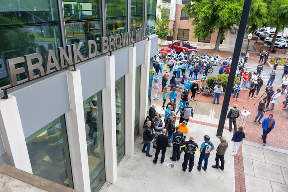 Outdoor event at Frank D. Brown Hall. Large letters on the side of the modern glass-and-concrete building identify the hall. A diverse crowd and a line of motorcycles are visible in the plaza and street below.