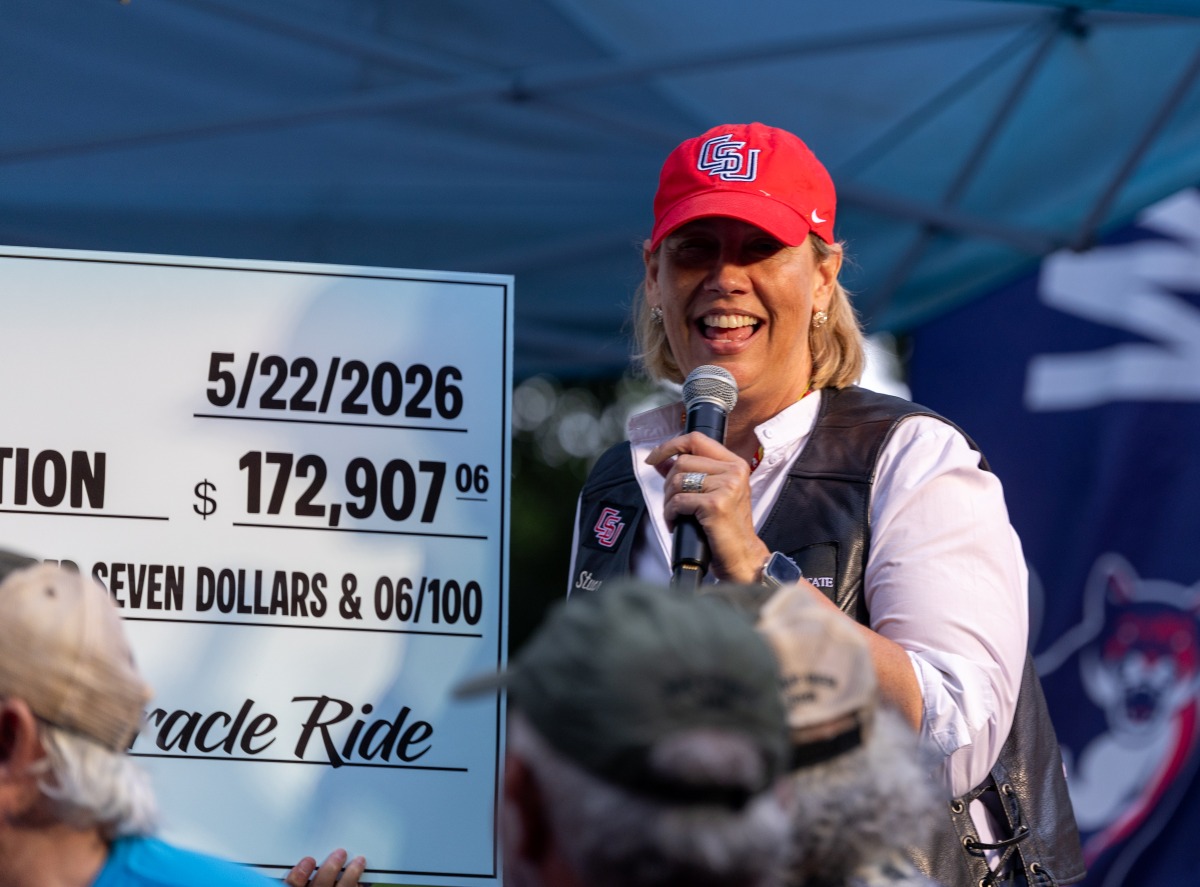 A smiling woman in a red CSU cap speaks into a microphone next to a large presentation check from the Miracle Ride dated 5/22/2026 for $172,907.06.