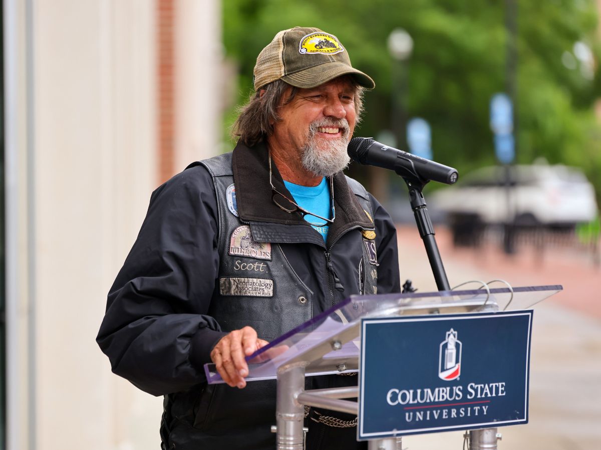 Close-up of a speaker at a Columbus State University event. The man, identified by a patch as 'Scott,' is wearing a leather motorcycle-style vest over a jacket and a cap. He is smiling into a microphone while standing behind a university-branded podium.