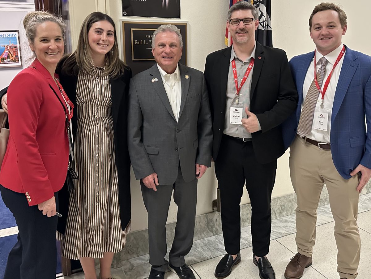 Five people stand in a line smiling for a photo indoors against a wall with a plaque that reads "REPRESENTATIVE Earl L. 'Buddy' Carter". From left to right: a woman in a red blazer and black pants; a woman in a brown and white striped dress with a black coat; a man in a grey suit; a man wearing glasses, a black suit jacket, and a name badge giving a thumbs-up; and a man in a blue blazer, khaki pants, and a tie.