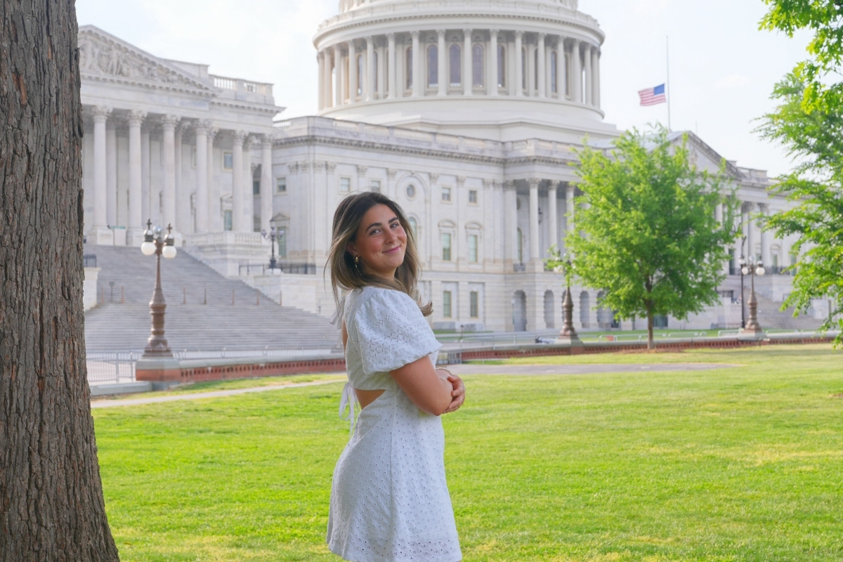 A young woman with long brown hair smiles over her shoulder while standing on a grassy lawn. She is wearing a short white eyelet dress with an open back. In the background, the United States Capitol dome and building are visible under a bright sky, partially framed by a large tree trunk on the left.