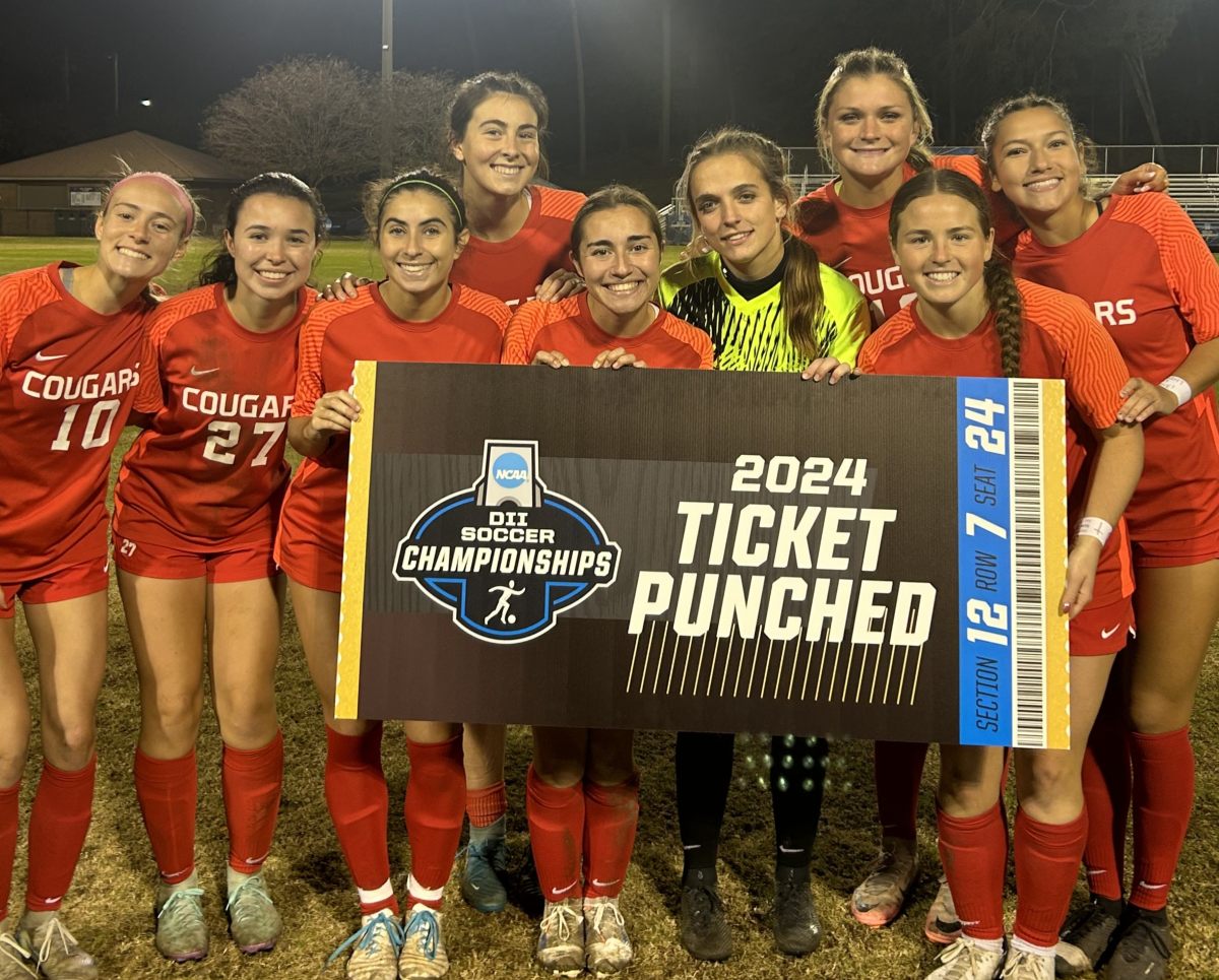 A group of female soccer players smile and pose together on a field at night under stadium lights. Most are wearing red jerseys that say "COUGARS," while one wears a yellow goalkeeper jersey. They are holding up a large sign designed to look like a ticket that reads, "NCAA DII SOCCER CHAMPIONSHIPS, 2024 TICKET PUNCHED, SECTION 12 ROW 7 SEAT 24."