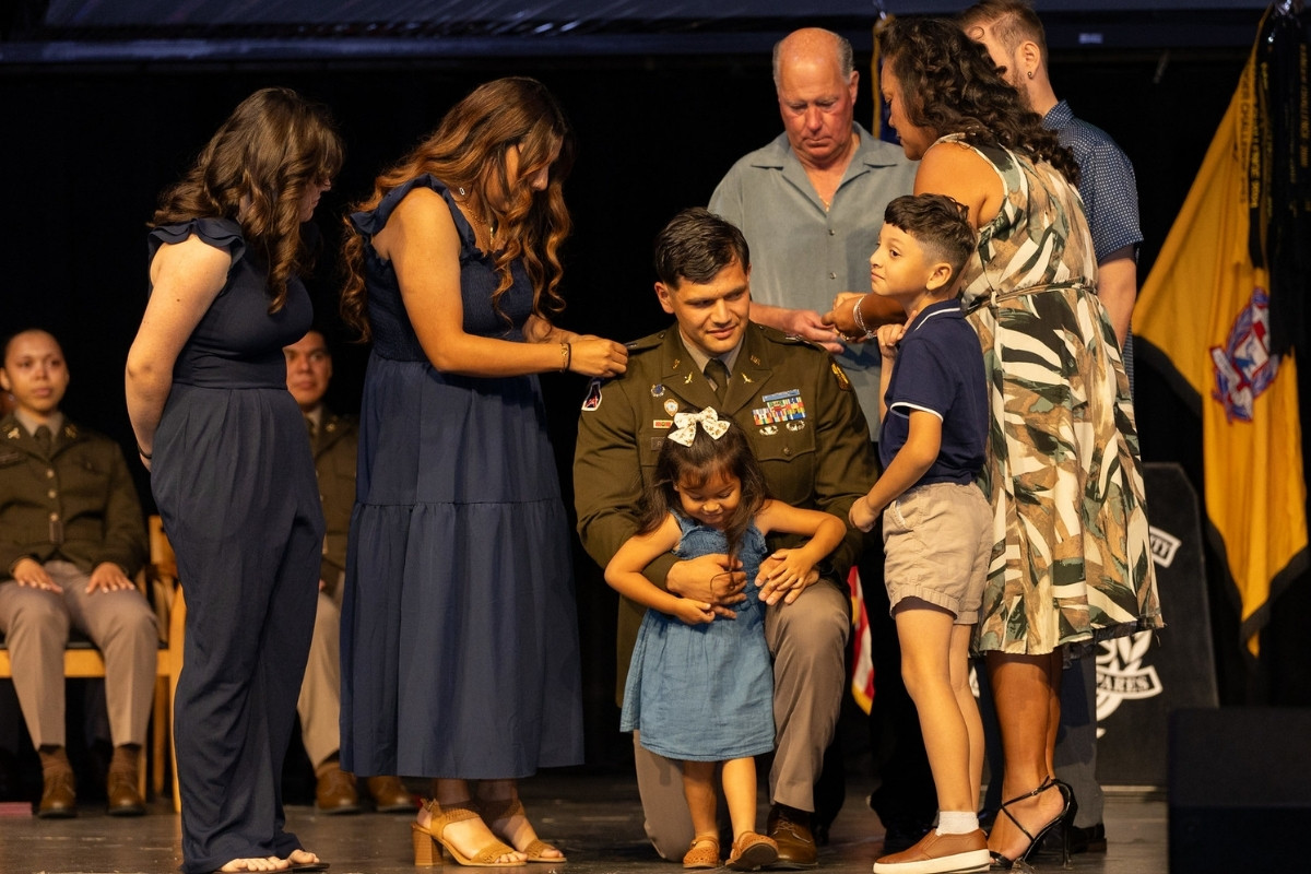 A military officer in a green service uniform kneels on a stage, surrounded by his family during a promotion or pinning ceremony. A woman in a blue dress adjusts the insignia on his shoulder, while a young girl stands in front of him and a young boy stands to the right. Other family members and military personnel look on in a dimly lit auditorium setting.