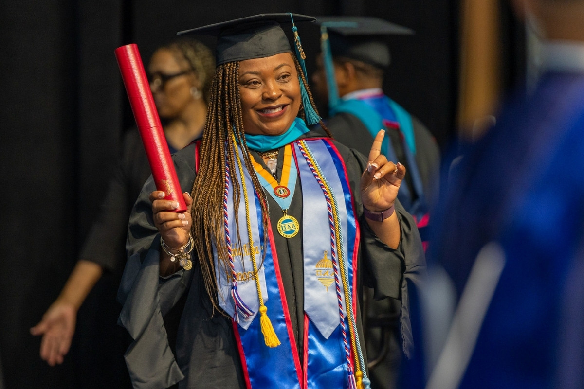 A smiling female graduate with long braids stands on a stage during a commencement ceremony. She is wearing a black cap and gown, a bright blue stole with red trim, a light blue honors stole, and multiple graduation cords and medals, including a circular gold 