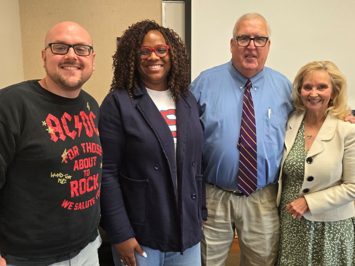 Four people stand shoulder-to-shoulder smiling for a photo indoors against a plain wall. From left to right: a man with a shaved head and glasses wearing a black AC/DC "For Those About to Rock" sweatshirt; a woman with curly hair and red glasses wearing a navy blazer over a graphic shirt; a man wearing glasses, a light blue button-down shirt, a striped tie, and khaki pants; and a woman with blonde hair wearing a white jacket over a green and white patterned dress.