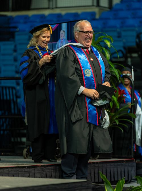 A man and a woman in academic regalia laugh during a graduation hooding ceremony on a stage. The woman, standing behind the man, places a master's hood over his shoulders. Both are wearing black gowns with blue accents, and the man wears a blue stole with red trim. Empty blue stadium seating is visible in the background.