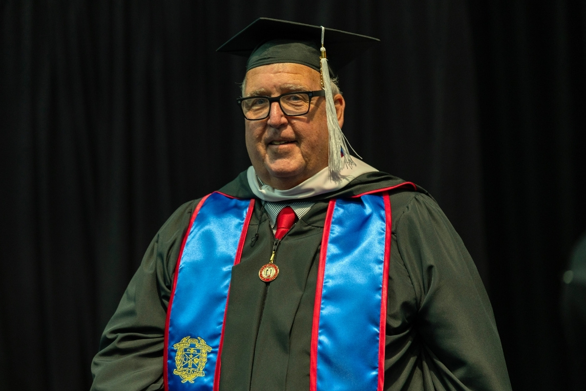 A medium close-up portrait of a man smiling in graduation regalia against a black curtain background. He is wearing glasses, a black mortarboard cap with a silver tassel, a black gown, a red tie with a small round medallion, and a bright blue stole with red trim featuring a gold embroidered crest on the lower left side.