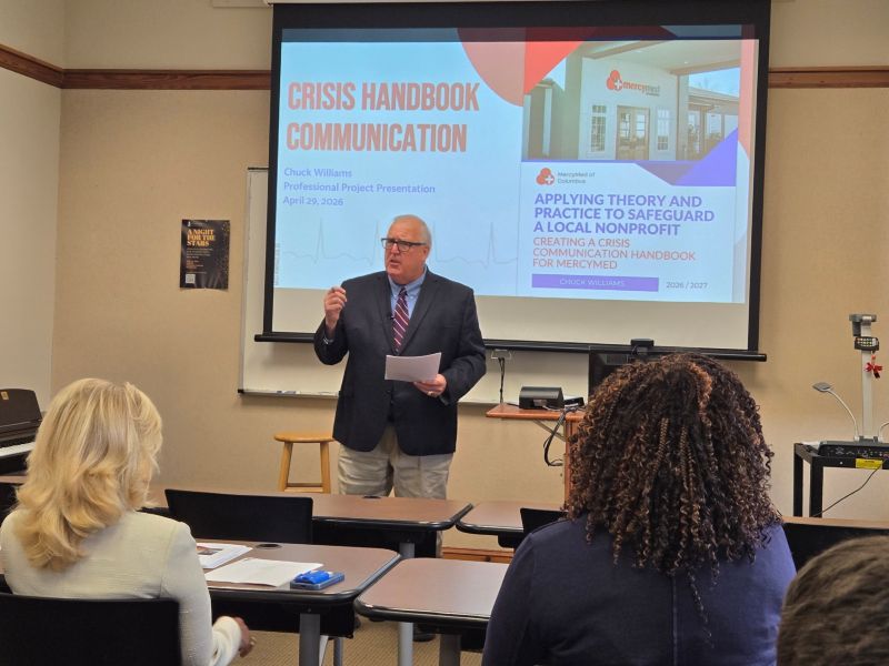 A man in a dark suit jacket, striped tie, and glasses stands at the front of a classroom presenting, holding a piece of paper and gesturing. Behind him, a projector screen displays a presentation slide titled "CRISIS HANDBOOK COMMUNICATION" by Chuck Williams, dated April 29, 2026, with a subtitle mentioning "MercyMed". In the foreground, the backs of two audience members—a blonde woman on the left and a woman with dark curly hair on the right—are visible listening to the presentation.