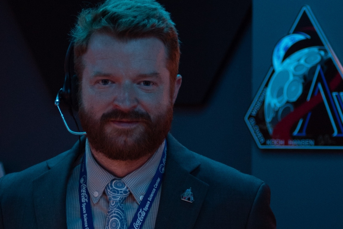 A close-up of a bearded man in a suit wearing a headset and a lanyard that reads "Coca-Cola Space Science Center." He is looking into the camera under dim blue lighting, with a stylized space mission emblem visible on the wall behind him.