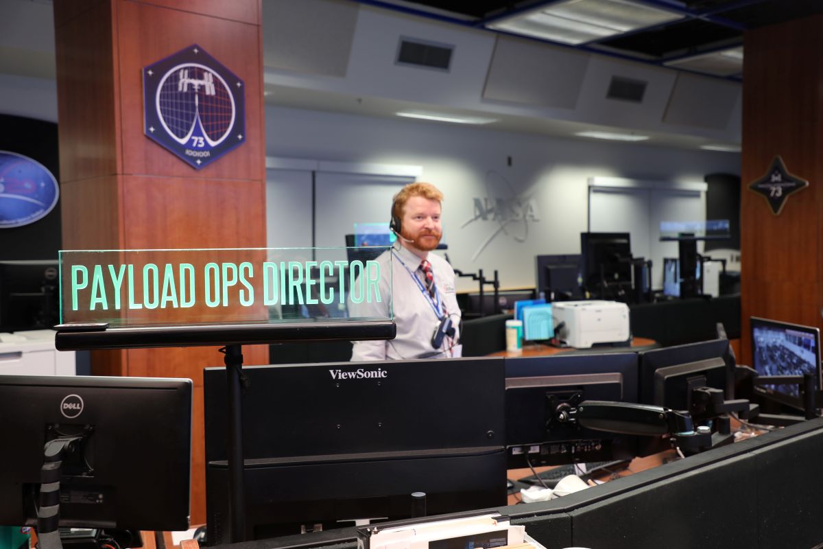 A man with red hair wearing a headset stands in a mission control center behind a row of computer monitors. In the foreground, a glowing glass sign reads "PAYLOAD OPS DIRECTOR," and a NASA logo is visible on the far wall in the background.