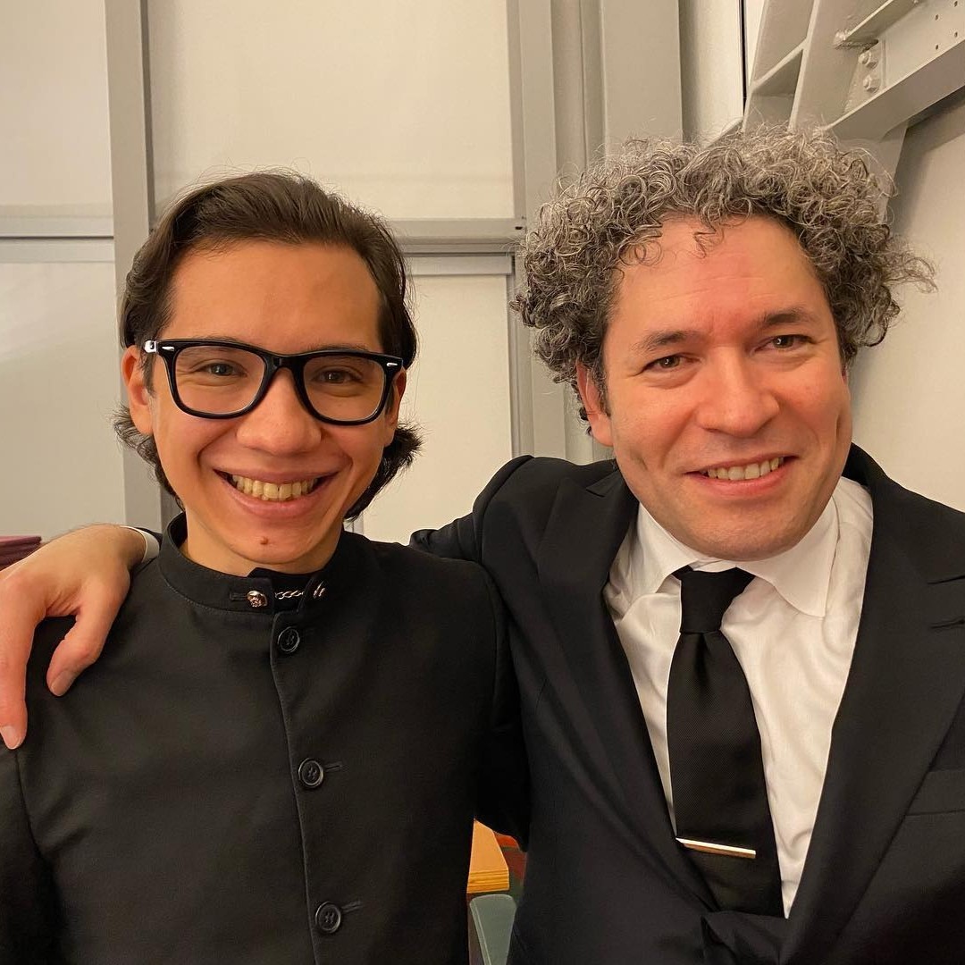 Two smiling men in formal black attire pose for a close-up photo together indoors.