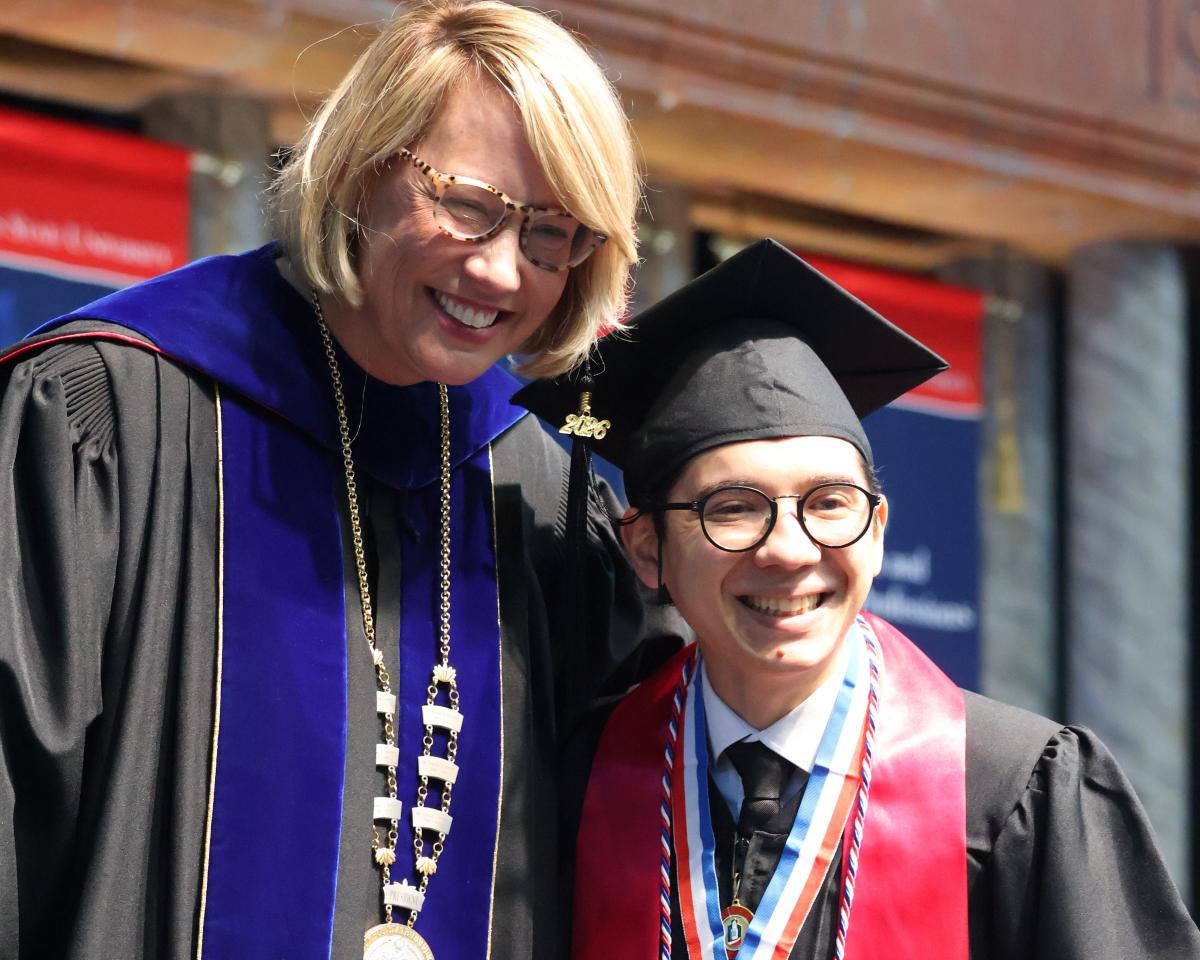 A university president in doctoral regalia poses with a smiling male graduate wearing a 2026 graduation cap, red stole, and honors medal.