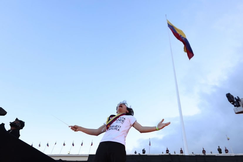 A low-angle shot of a musical conductor looking upward on an outdoor stage beneath a large Venezuelan flag flying high on a flagpole.