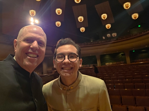 Two smiling men take a selfie inside an empty theater auditorium with rows of red seats and distinct, glowing modern ceiling lights.