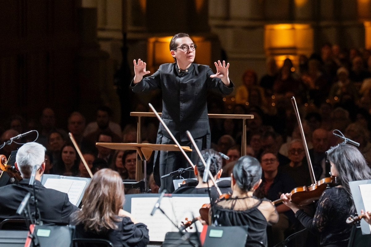 An orchestral conductor gestures dynamically while leading a performance, viewed from behind the musicians.