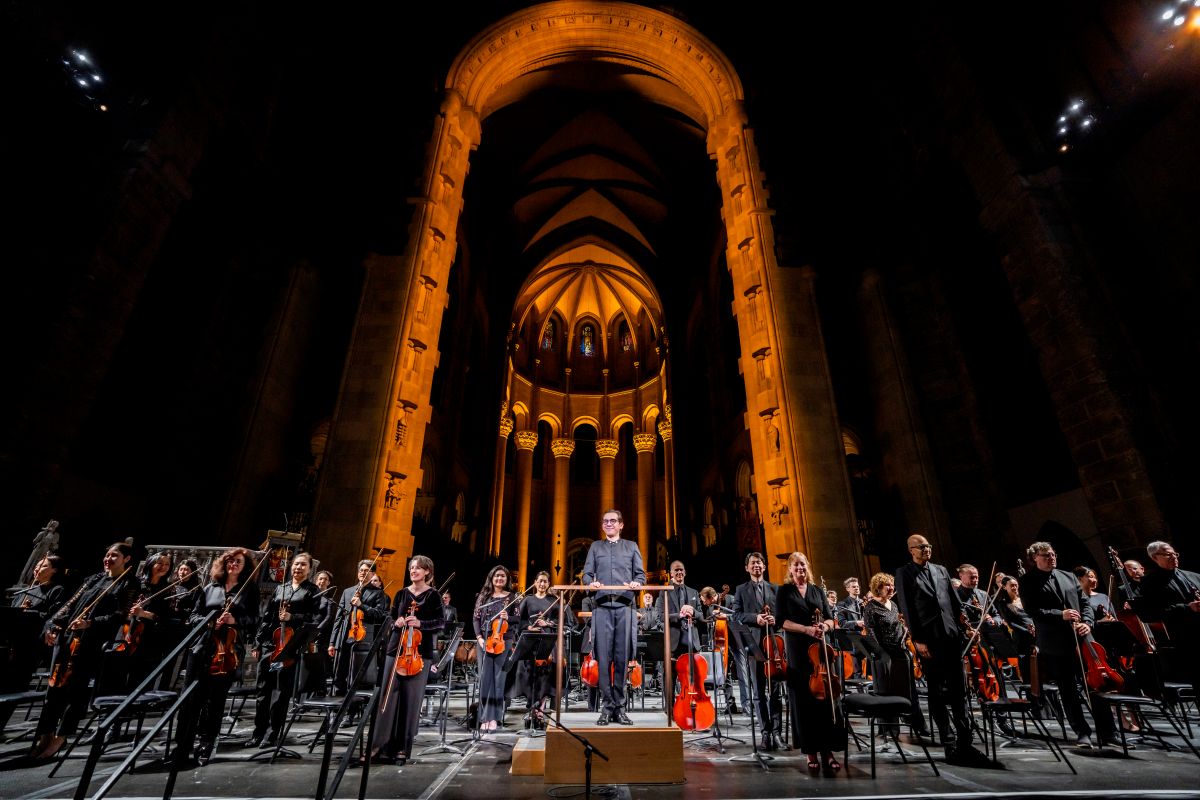 A large orchestra and their conductor stand on stage during a performance inside a massive, warmly lit gothic cathedral.