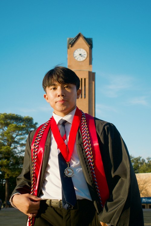 A young man in a black graduation gown, tie, red stole, braided honor cords, and a medal poses outdoors on a sunny day. A tall brick clock tower stands prominently in the background against a clear blue sky.
