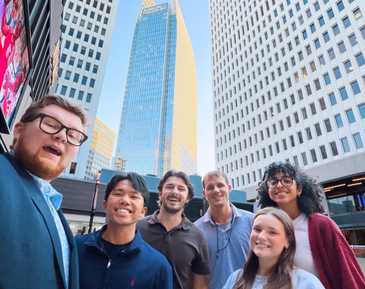 A group of six smiling young adults poses for a low-angle selfie outdoors in a city center. Towering high in the background are major skyscrapers, including a prominent curved glass building with "King & Spalding" visible near the top, flanked by a tall white high-rise under a clear blue sky.