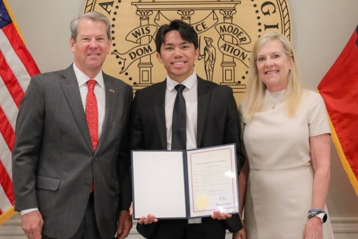 A young man in a black suit and tie stands between Georgia Governor Brian Kemp and First Lady Marty Kemp while holding an open certificate folder. They are posing in front of a large, circular gold seal of the State of Georgia, flanked by the United States and Georgia state flags.
