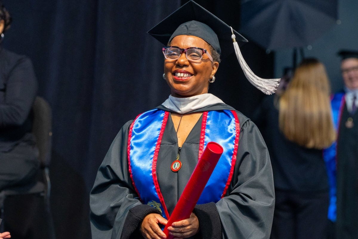 A smiling woman wearing a black graduation cap with a white tassel, glasses, and an academic gown with a bright blue and red stole looks forward while holding a red diploma tube. She is standing on a stage with a dark curtain backdrop and other graduates in the background.