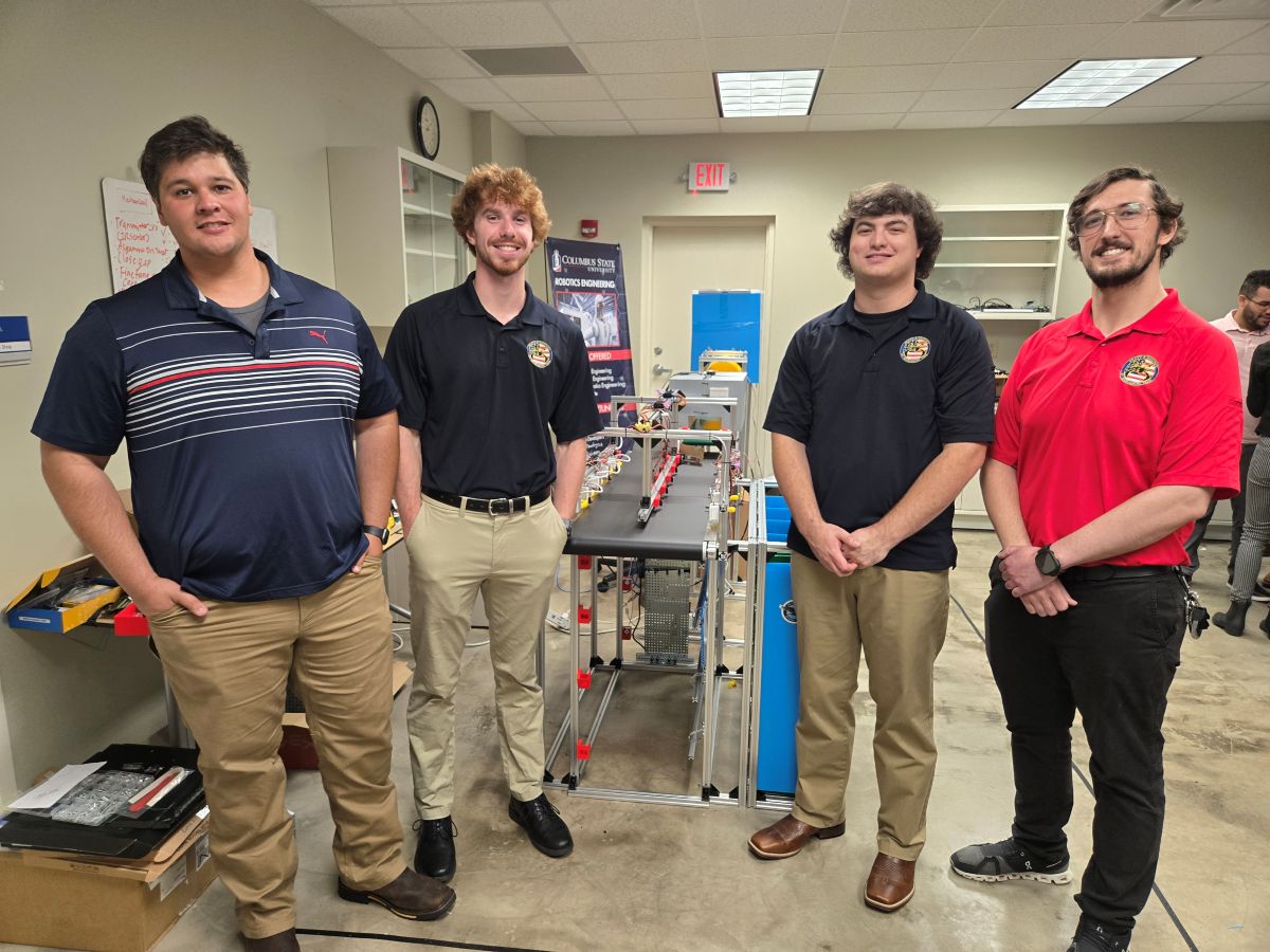 group portrait of four Columbus State University robotics engineering students standing with their automated machinery capstone project inside a university laboratory.