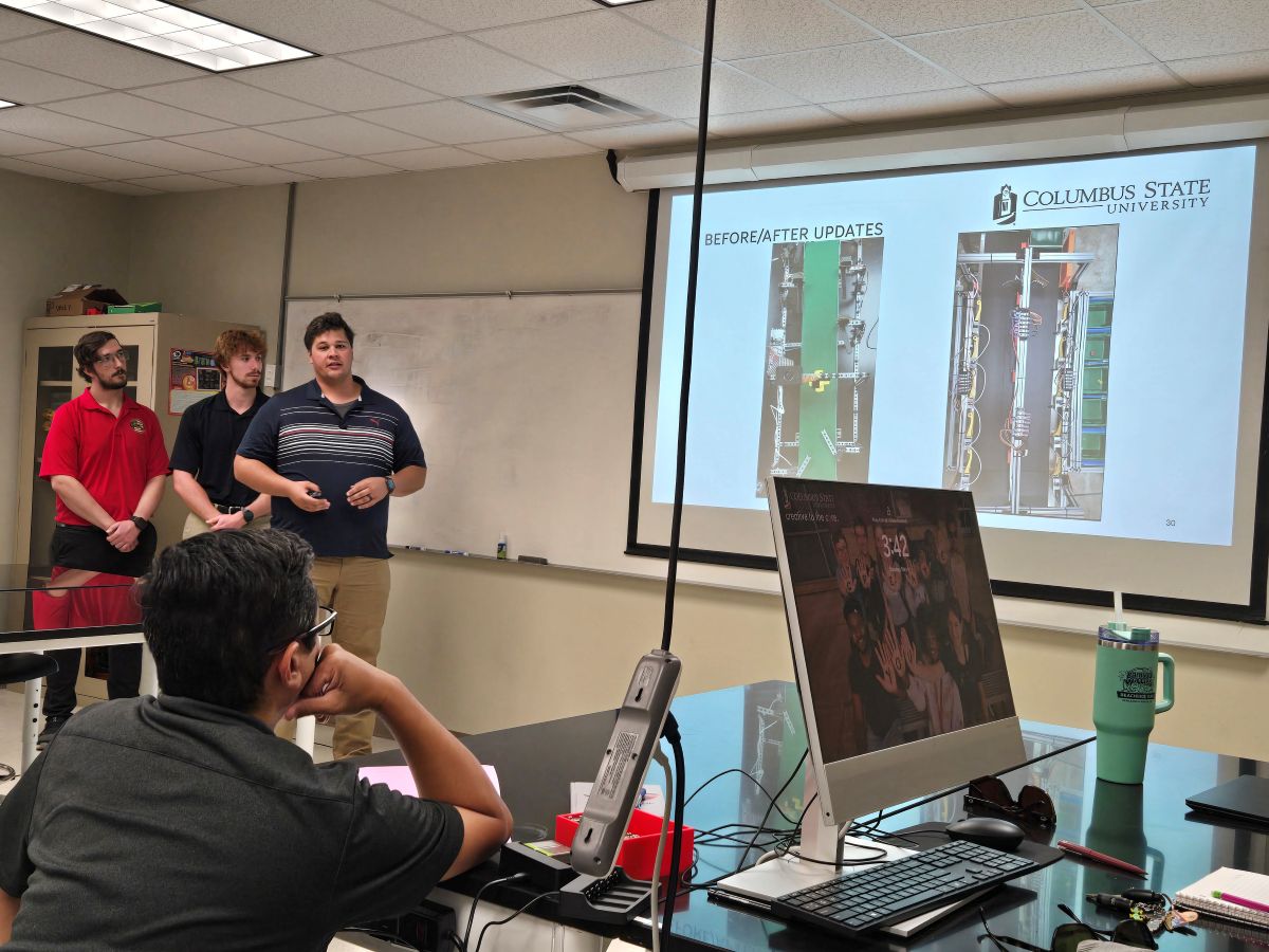 Columbus State University engineering students give a presentation on mechanical design updates using a projector slide, while an instructor or classmate listens from a laboratory workspace.