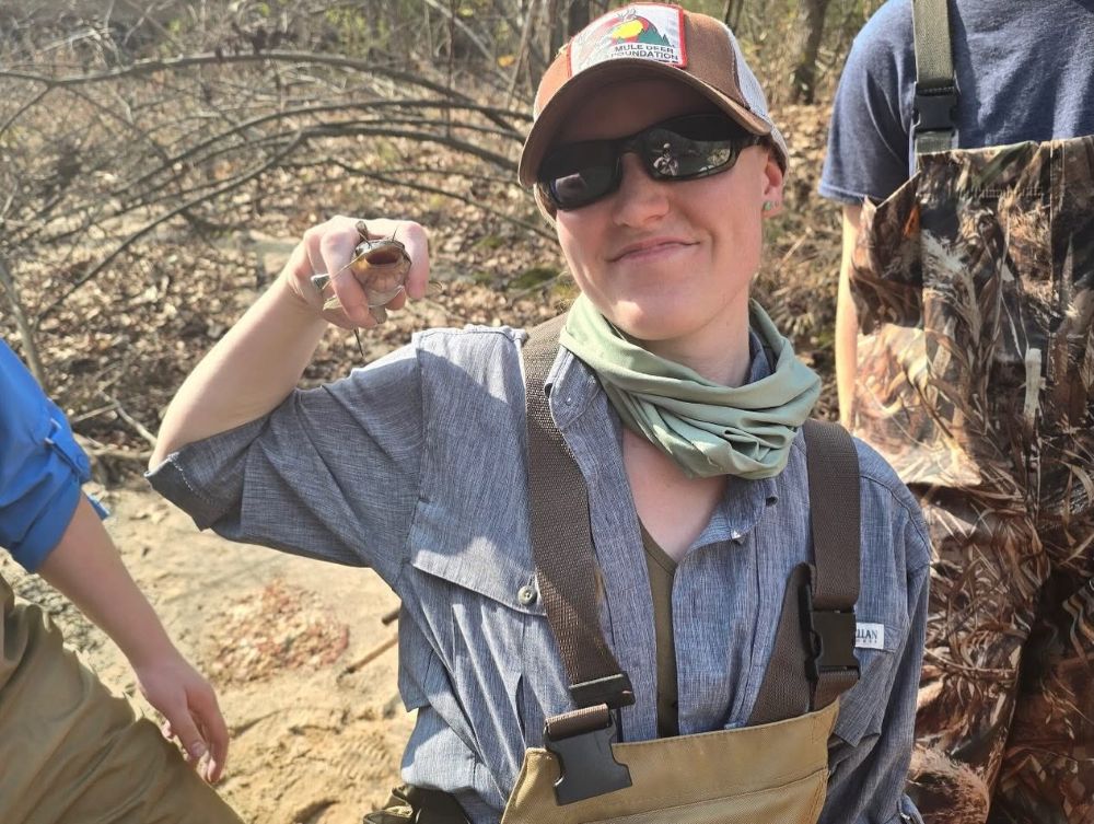 A close-up of a person wearing a brown Mule Deer Foundation cap, sunglasses, a green neck gaiter, and fishing waders. They are smiling slightly while holding up a small catfish for the camera outdoors in a sunny, wooded environment.