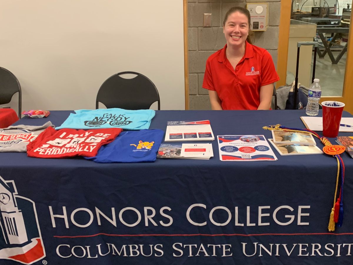 Kaylee Dodson sitting behind a table with a Columbus State University Honors College tablecloth and paraphernalia on the table.