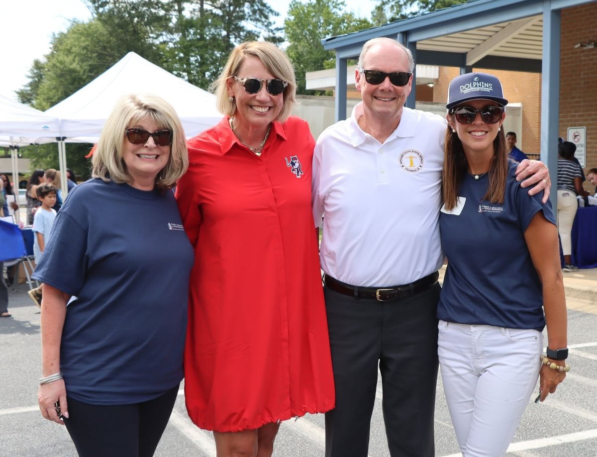 Four smiling adults standing together outdoors in a parking lot setup for an event. From left to right, a woman in a navy shirt and sunglasses, a taller woman in a bright red shirt dress, a man in a white polo shirt, and a woman wearing a navy "Dolphins" cap, navy shirt, and white pants.