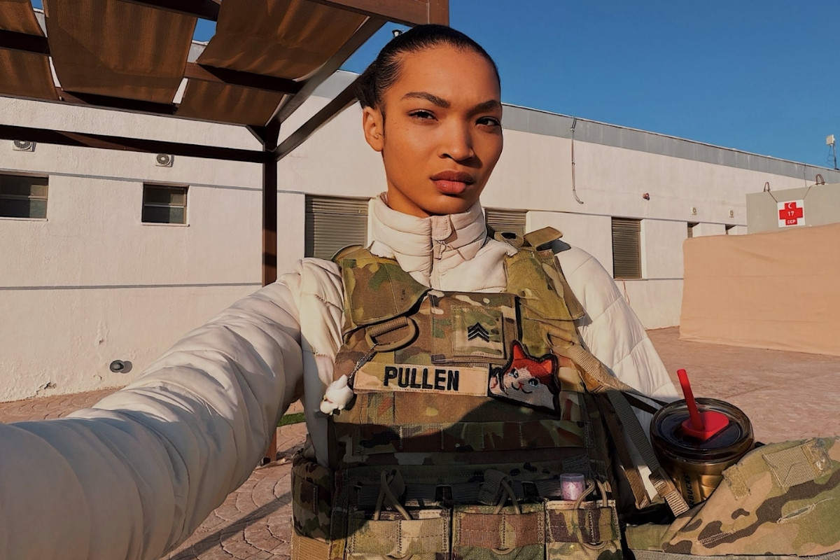 A woman in military gear takes a selfie outdoors in front of a white building. She is wearing a cream-colored puffer jacket under a camouflage tactical vest that features a name tape reading "PULLEN," a Sergeant rank insignia, and a cartoon cat patch.
